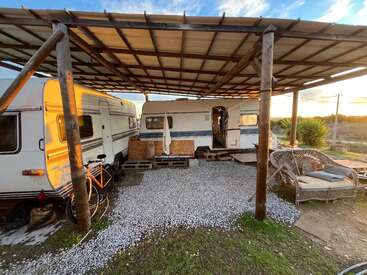 Two retro caravans sit under a rustic wooden canopy. There’s a bicycle outside, wicker sofa seating, gravel ground, and warm sunlight illuminating the peaceful outdoor setup.