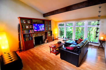 L'image représente un salon doté d'une grande fenêtre, avec un canapé, une table basse et une lampe, sur un mur de couleur neutre et un sol de couleur claire. La pièce est bien éclairée et aérée.