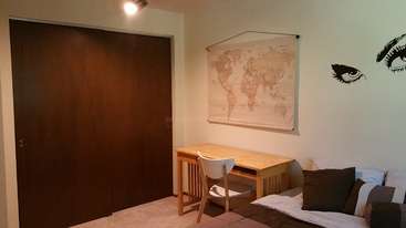 L'image représente une pièce avec une table et des chaises, dotée d'une grande fenêtre, d'une bibliothèque et d'un bureau, avec une personne assise à ce dernier, le tout dans un environnement de couleurs neutres.