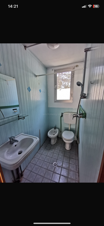A imagem mostra um banheiro com pia, vaso sanitário e banheira, com paredes e piso brancos e uma janela no lado esquerdo do cômodo.