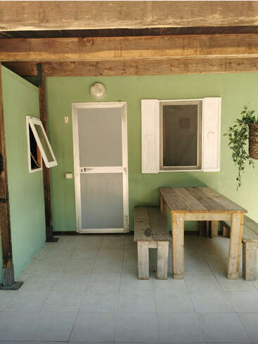 A imagem retrata um espaço externo sereno com uma mesa e bancos de madeira, tendo como pano de fundo paredes verdes e uma porta branca, evocando uma sensação de calma e tranquilidade.
