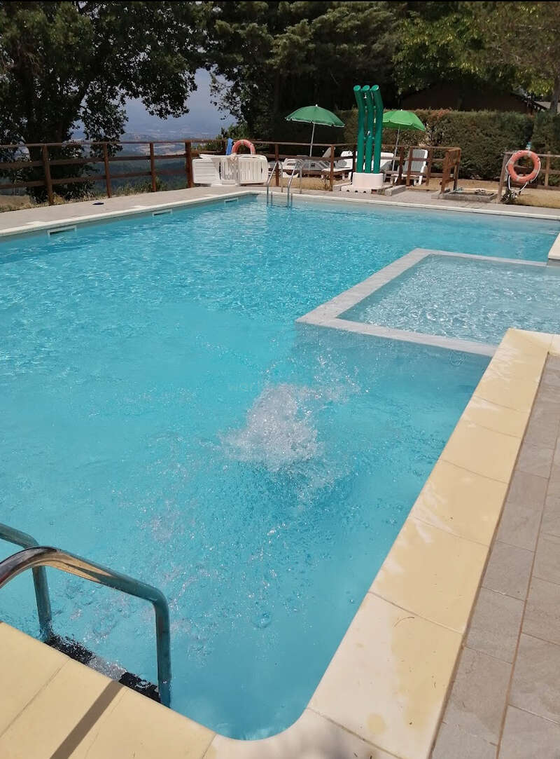 A imagem retrata uma cena serena de piscina externa, com uma piscina retangular com uma banheira de hidromassagem quadrada, cercada por espreguiçadeiras e guarda-sóis, em um cenário pitoresco com árvores.