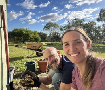 A smiling couple enjoys gardening outdoors on a sunny day. They are surrounded by pots, soil, and a wheelbarrow, with trees and blue sky behind them.