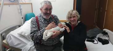 An elderly man and woman happily sit on a hospital bed, smiling and holding a newborn baby. The background includes hospital furnishings and personal items, creating a warm atmosphere.