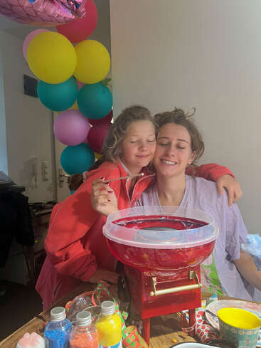 Deux jeunes filles souriantes sont assises l'une à côté de l'autre, les yeux fermés, profitant d'un moment de joie. Des ballons colorés et une machine à barbe à papa suggèrent une atmosphère de fête et de célébration.