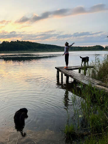 Une personne se tient sur un quai en bois au bord d'un lac au coucher du soleil, s'étirant joyeusement. Deux chiens se trouvent à proximité, l'un sur le quai, l'autre dans l'eau.