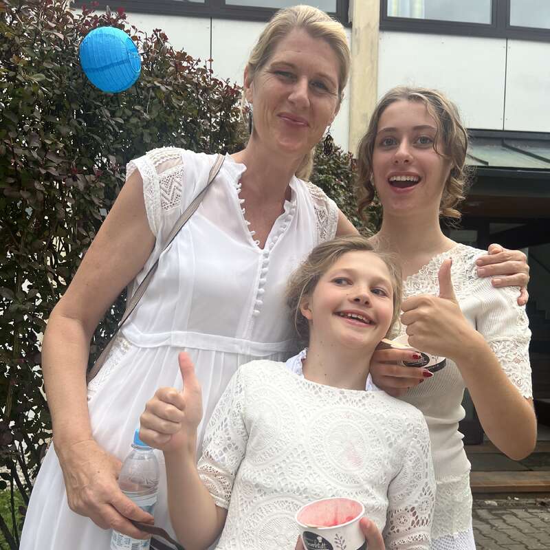 Une femme et deux filles, toutes vêtues de blanc, sourient à l'extérieur. Les filles montrent joyeusement leur pouce en l'air, tenant des coupes de crème glacée. Lanterne bleue en arrière-plan.