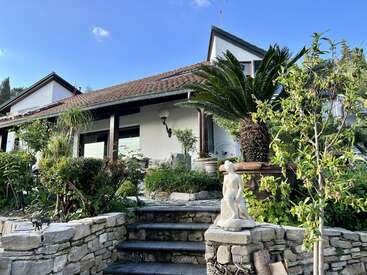 La imagen muestra una serena casa blanca con tejado marrón, rodeada de exuberante vegetación y una escalera de piedra que conduce a la entrada, en la que aparece la estatua de una persona.