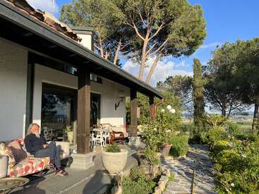 La imagen representa una serena escena de una mujer sentada en un porche, rodeada de exuberante vegetación y un paisaje pintoresco, que destila una sensación de tranquilidad y relajación.