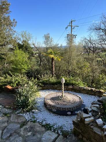 La imagen representa un sereno jardín con un patio de piedra, guijarros blancos y una singular escultura, sobre un fondo de árboles y un cielo azul despejado.
