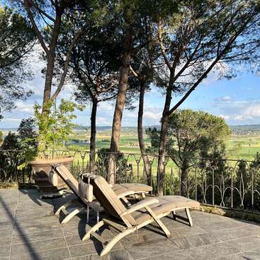 Das Bild zeigt eine ruhige Terrassenszene mit zwei Liegestühlen, einem Topfbaum und einem schmiedeeisernen Zaun mit Blick auf eine malerische Landschaft mit Bäumen und Feldern unter einem blauen Himmel.
