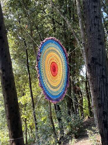 Uma mandala de crochê grande e colorida está suspensa entre as árvores em uma floresta, criando uma exibição de arte circular vibrante, cercada por vegetação e luz natural.