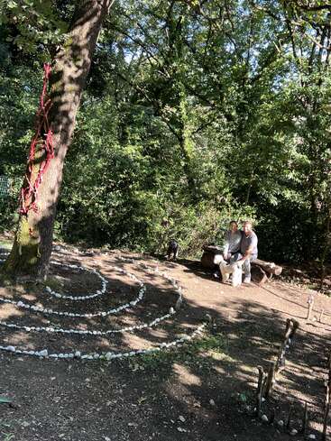 Um casal senta-se em um banco rústico em uma área arborizada tranquila, cercado por círculos concêntricos de pedra. Dois cães estão por perto. A luz do sol passa por entre as árvores.
