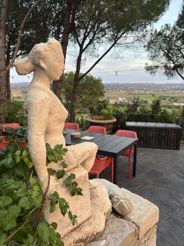 Uma estátua de pedra envelhecida de uma mulher fica em uma saliência, com vista para mesas e cadeiras ao ar livre, cercada por vegetação, com colinas panorâmicas ao fundo.