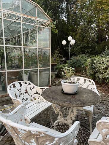 Um ambiente externo encantador, com cadeiras brancas ornamentadas e uma mesa redonda de pedra com um vaso de plantas, ao lado de uma estufa de vidro cercada por uma vegetação exuberante.