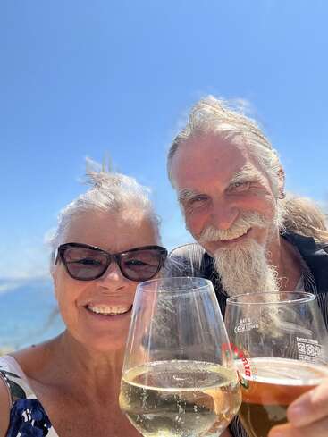 Um casal de idosos sorridente desfruta de um dia ensolarado ao ar livre, segurando copos de vinho branco e cerveja, comemorando juntos com o céu azul e a água ao fundo.