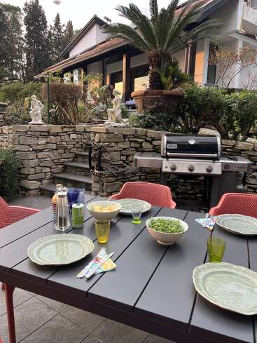 A beautifully set outdoor dining table with plates, drinks, and bowls of food. There’s a grill, stone wall, garden statues, lush greenery, and a charming house.