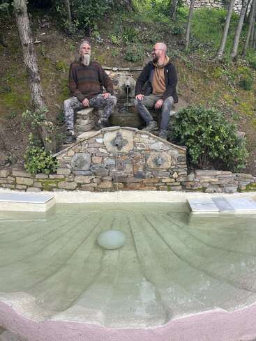Two men sit on a stone fountain surrounded by greenery. One has a beard and sweater, the other wears glasses and a hoodie; both look relaxed.