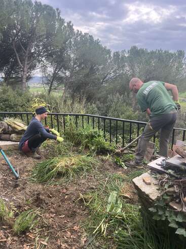 Two people are gardening on a hillside, removing weeds and grass. Surrounded by trees and cloudy sky, they work near a black fence with scattered tools nearby.