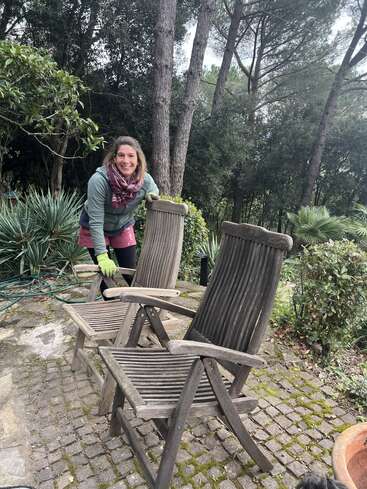 A smiling woman in outdoor clothing and gloves stands on a stone patio with two wooden chairs, surrounded by lush green foliage and tall trees.