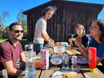 Vier Freunde genießen ein sonniges Essen im Freien, lächeln und essen Pizza an einem Holztisch, umgeben von Getränken und Essen, vor einem rustikalen Holzschuppen.