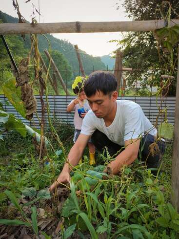 Um homem está ajoelhado em um jardim, cuidando das plantas. Uma criança está atrás dele, bebendo de uma bolsa, cercada por vegetação e montanhas. Cena tranquila.