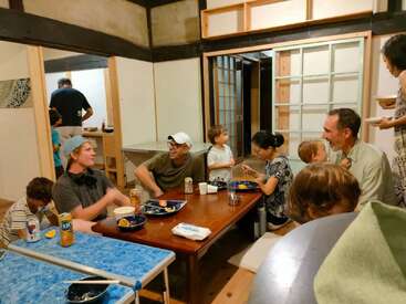 Um grupo de pessoas, adultos e crianças, desfruta de uma refeição casual em uma sala de jantar aconchegante, rindo, conversando e compartilhando alimentos, criando uma atmosfera calorosa.