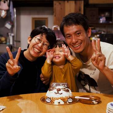 Uma família alegre de três pessoas posa junta, sorrindo e fazendo sinais de paz. Um lindo bolo com tema de animais está na frente deles, comemorando uma ocasião especial.
