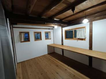 Esse quarto tem piso de madeira, vigas expostas no teto, paredes brancas, três janelas pequenas, uma janela maior com cortinas e uma longa mesa de madeira em uma das paredes.