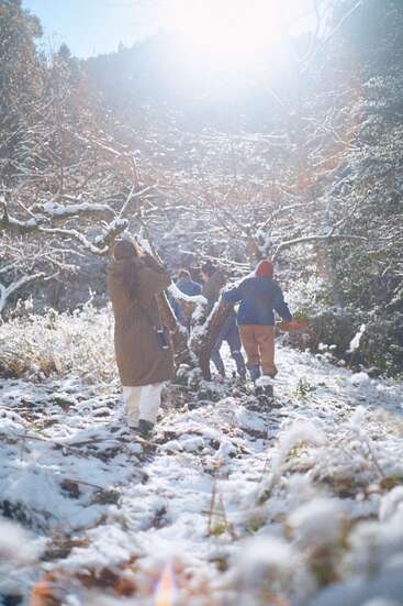 Um grupo de pessoas caminha por uma trilha na floresta nevada sob a luz do sol, vestidas com casacos e chapéus de inverno, cercadas por árvores congeladas e pela natureza.