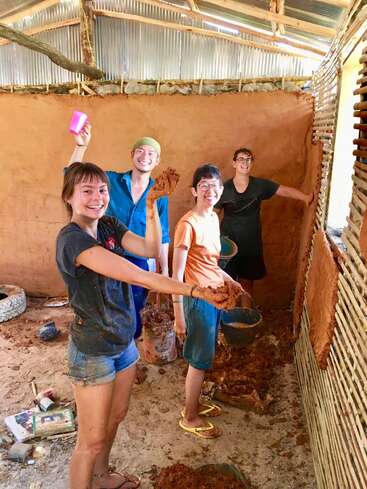 Quatro jovens sorrindo enquanto constroem uma parede de barro em uma estrutura rústica. Eles estão descalços, se divertindo e usando suas mãos e baldes para a construção.