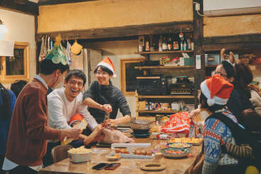 Um grupo de amigos e familiares se reúnem em uma cozinha aconchegante, usando chapéus festivos, rindo e preparando juntos uma refeição de fim de ano em meio a decorações calorosas e alegres.