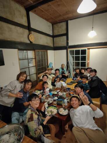 Um grande grupo de pessoas, incluindo crianças, se reúne em torno de uma mesa cheia de comida, sorrindo e fazendo sinais de paz em uma sala japonesa tradicional e aconchegante.