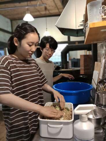 Duas pessoas estão em uma cozinha preparando comida juntas. Uma pessoa está misturando ingredientes em um recipiente enquanto a outra observa e sorri. Atmosfera aconchegante, luzes no teto.