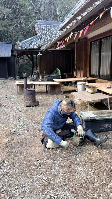 Une personne vêtue d'une veste bleue répare une tronçonneuse à l'extérieur d'une maison rustique au toit de tuiles, entourée d'arbres et de mobilier d'extérieur, avec des banderoles au-dessus. Cadre rural paisible.