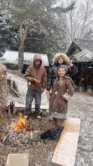 Trois personnes vêtues de manteaux d'hiver se tiennent près d'un feu de camp en plein air, entourées de neige qui tombe et de bâtiments rustiques. L'une d'elles fait le signe de la paix ; tout le monde semble bien au chaud.