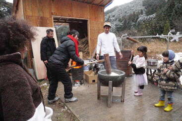 Un groupe de personnes, dont des enfants, prépare des mochi en plein air. Ils utilisent des maillets en bois et un grand mortier en pierre, entourés d'un paysage hivernal et d'un abri en bois.