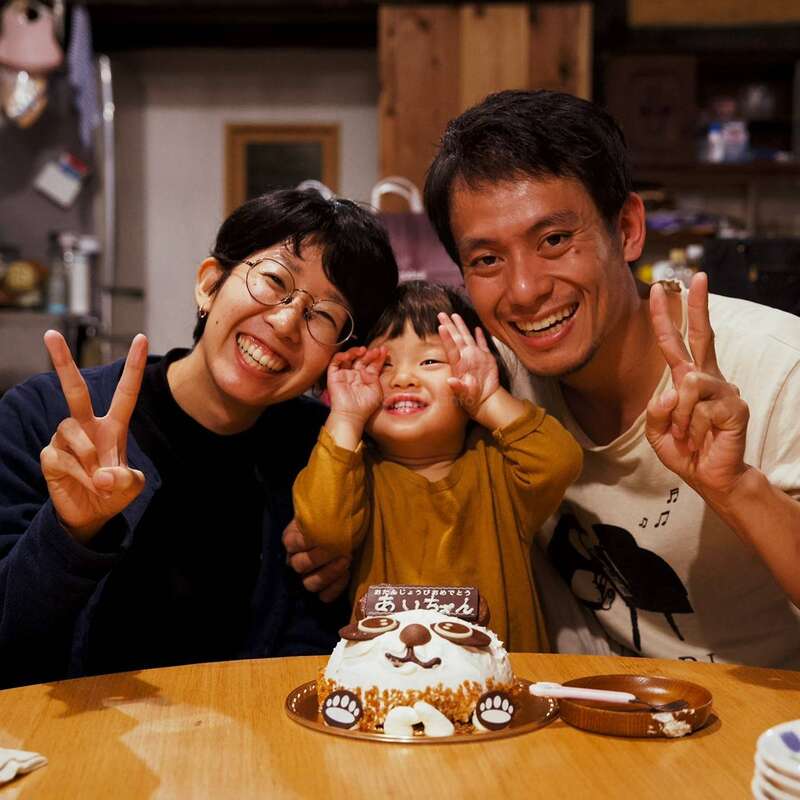 Uma família alegre de três pessoas posa junta, sorrindo e fazendo sinais de paz. Um lindo bolo com tema de animais está na frente deles, comemorando uma ocasião especial.