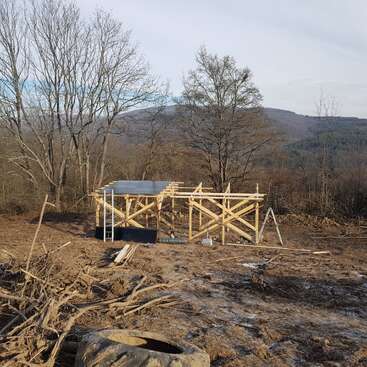 Une structure en bois est en cours de construction dans un champ aride et boueux, entouré d'arbres dépouillés et de collines, avec des matériaux de construction et des débris éparpillés tout autour.