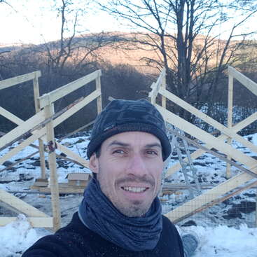 The image depicts a person constructing a wooden structure, likely a fence or gate, with a pile of lumber and tools nearby, set against a natural outdoor backdrop.