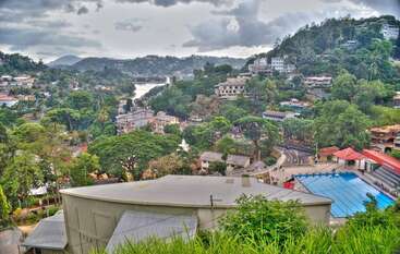 Una vibrante ciudad enclavada entre exuberantes colinas verdes, rodeada de árboles y edificios, con un río serpenteante y un estanque azul bajo un espectacular cielo nublado.