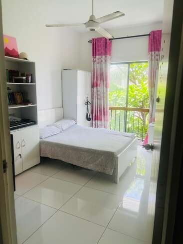 Un dormitorio luminoso y ordenado con suelo de baldosas blancas, una cómoda cama, estanterías, ventilador de techo, armario blanco, cortinas rosas y una gran ventana con vistas a los verdes árboles del exterior.