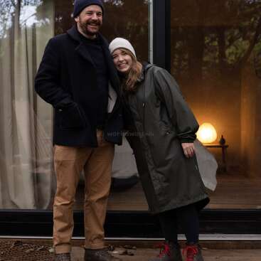 The image depicts a man and woman standing outside a glass door, with the woman wearing a long coat and the man in a dark jacket and hat.