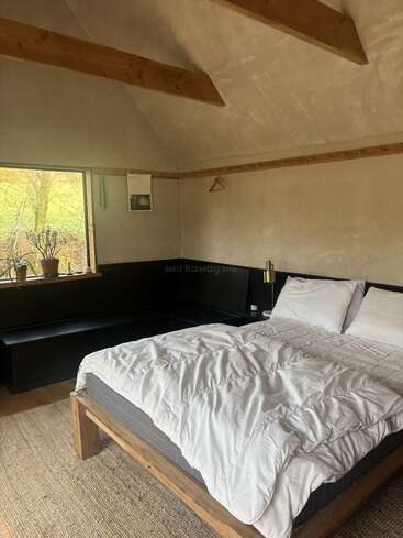 Este acogedor dormitorio cuenta con una cama de madera con ropa de cama blanca, vigas a la vista, un gran ventanal con vistas panorámicas, una planta en maceta, un banco y una decoración minimalista.
