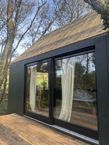 Una cabaña moderna con grandes puertas acristaladas y cortinas blancas, enclavada entre árboles desnudos. En la parte delantera hay una terraza de madera que se integra armoniosamente en el entorno natural y sereno.