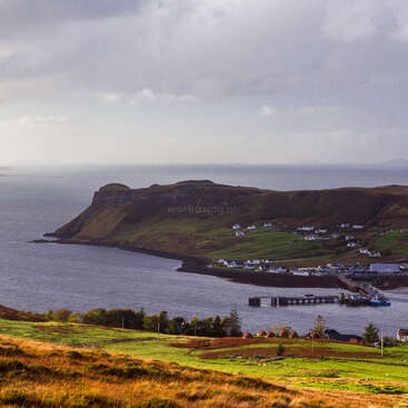 The image depicts a serene coastal landscape with a body of water, a pier, and a small town nestled among rolling hills under a cloudy sky.