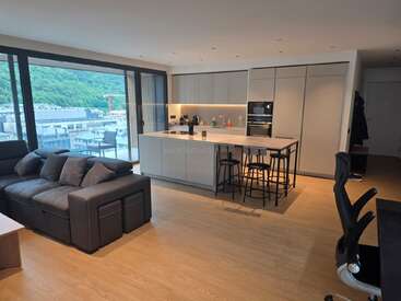 Apartamento moderno em plano aberto com uma cozinha elegante, ilha e bancos de bar, um aconchegante sofá modular cinza, janelas amplas com vistas panorâmicas e piso de madeira clara por todo o apartamento.