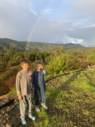 Two children stand on a grassy hill, smiling, with mountains and lush greenery behind them. A faint rainbow stretches across the cloudy sky in the background.