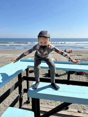 A child dressed in a wetsuit and cap balances playfully on a turquoise wooden lifeguard stand at the beach, with the ocean and people behind.