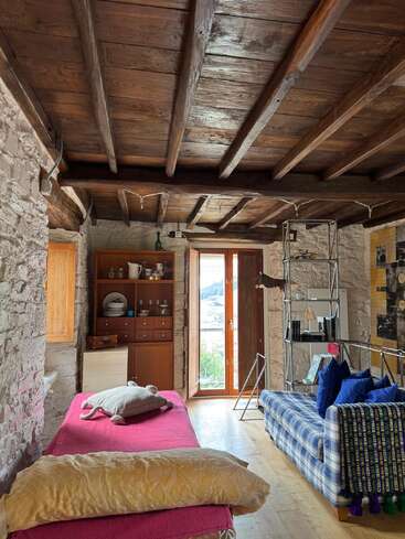 Esse quarto aconchegante tem paredes de pedra rústica, teto com vigas de madeira, um sofá xadrez com almofadas azuis, uma cama coberta de rosa e uma porta aberta com vistas panorâmicas.